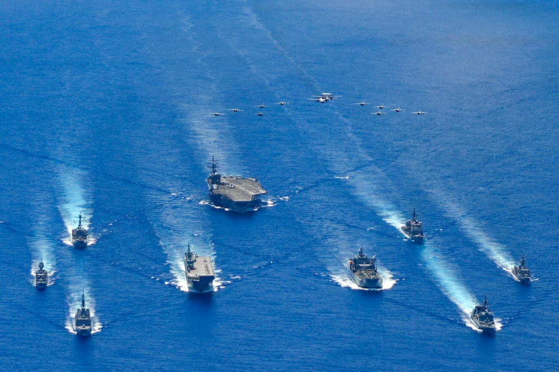 A large group of military ships move across the ocean in formation.