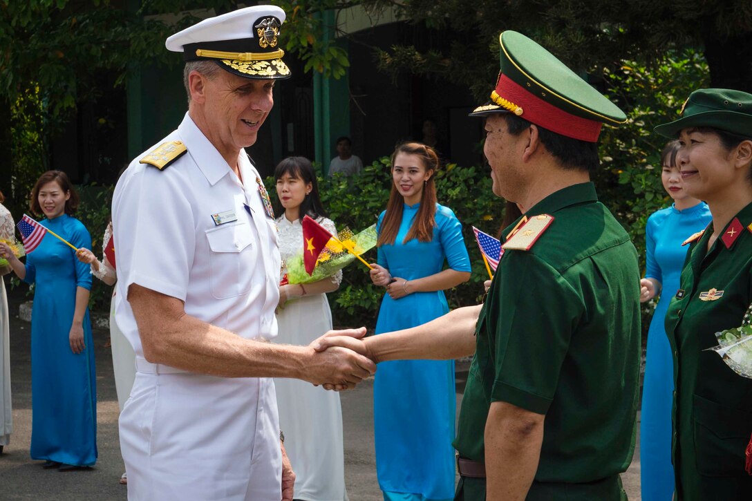 Two men dressed in military uniforms shake hands as others look on.
