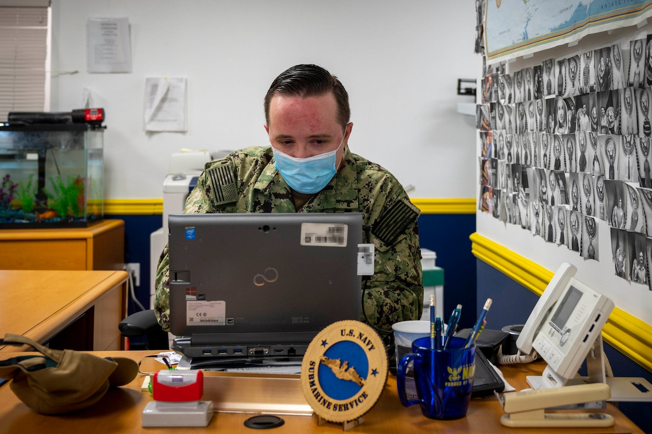 A sailor wearing a face mask works on a laptop.