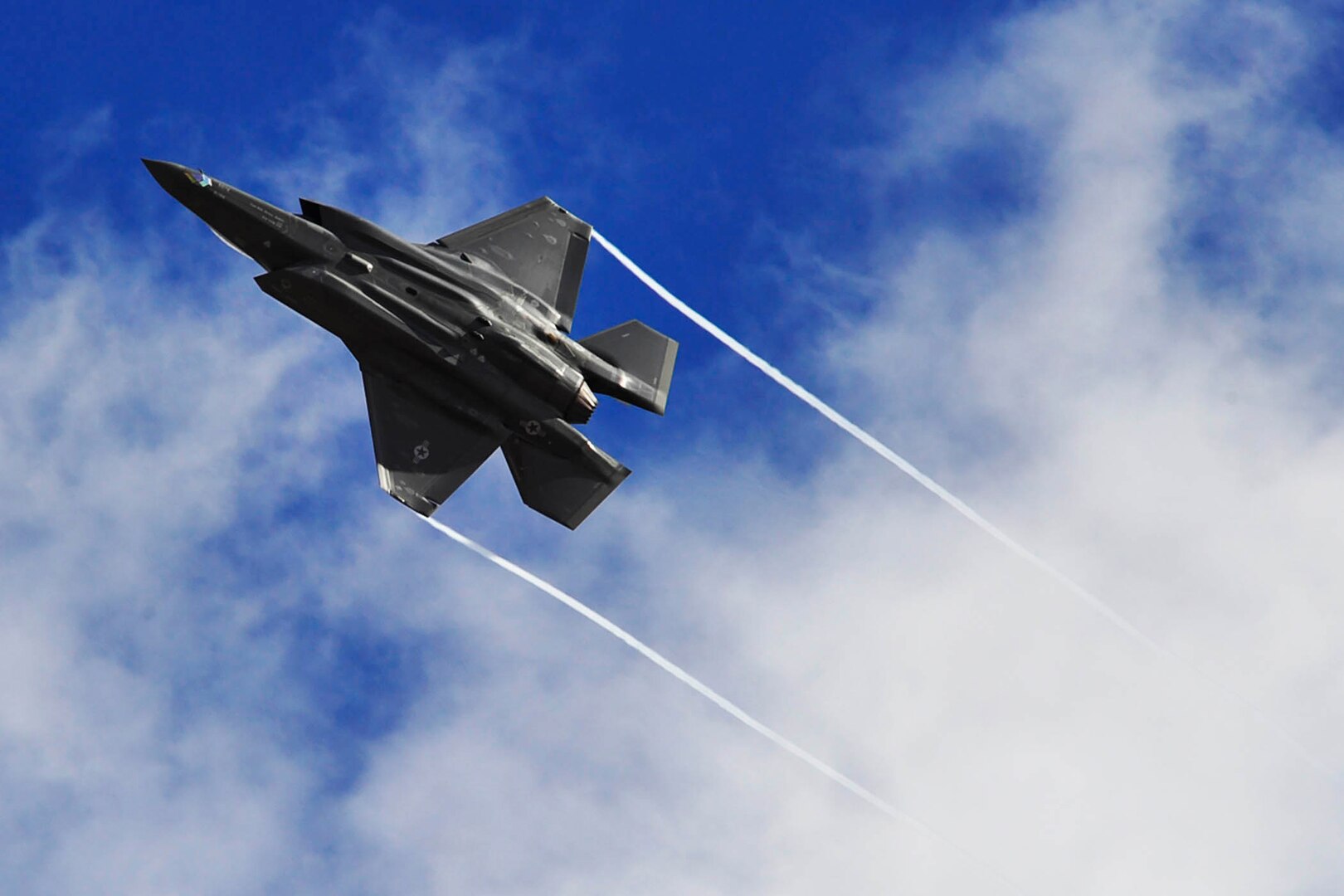 A fighter jet flies against clouds and a deep blue sky. Condensation trails from its wing tips.