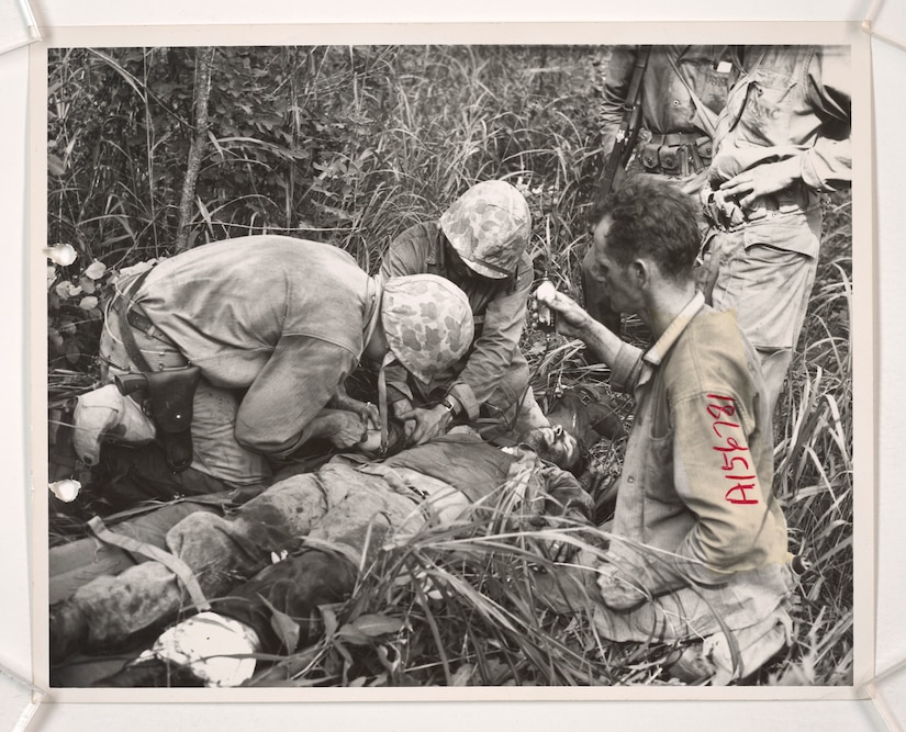 Three men in military uniforms work on an injured serviceman lying down in tall grass.