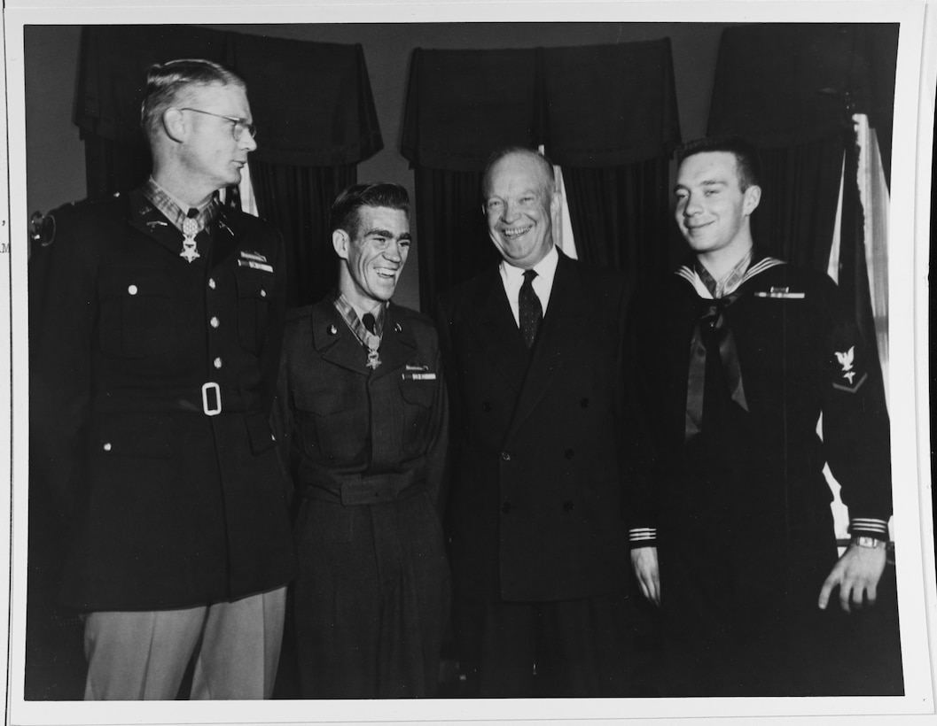 Four men, three in uniforms and wearing medals stand smiling.