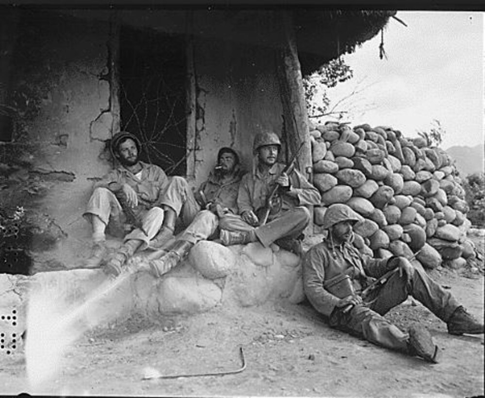Four men holding rifles rest beside a hut.