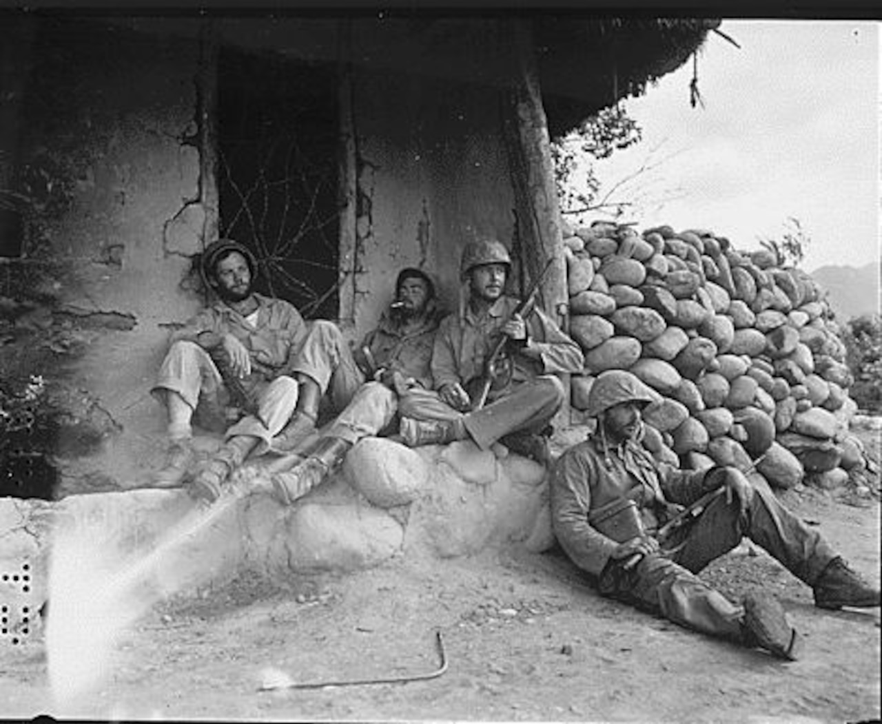 Four men holding rifles rest beside a hut.