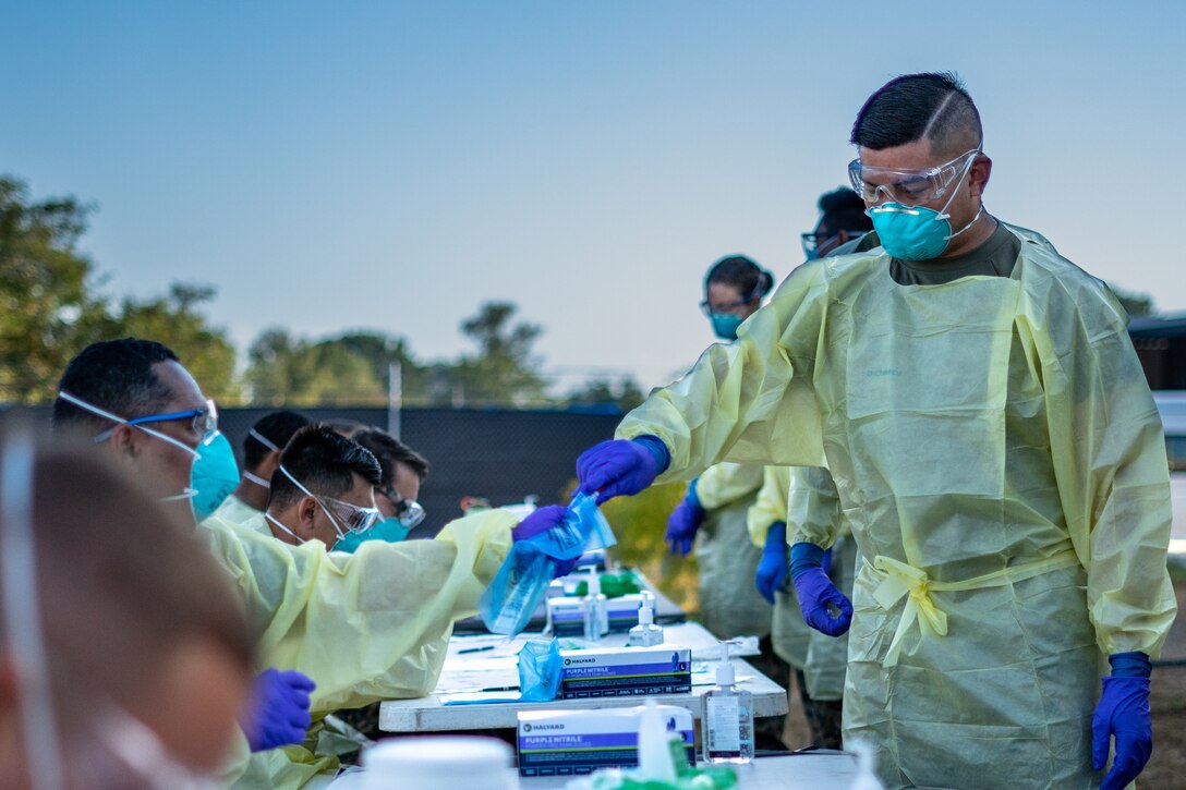 A group of people wearing personal protective equipment walk in a line in front of a group of people sitting at a table also wearing personal protective equipment.