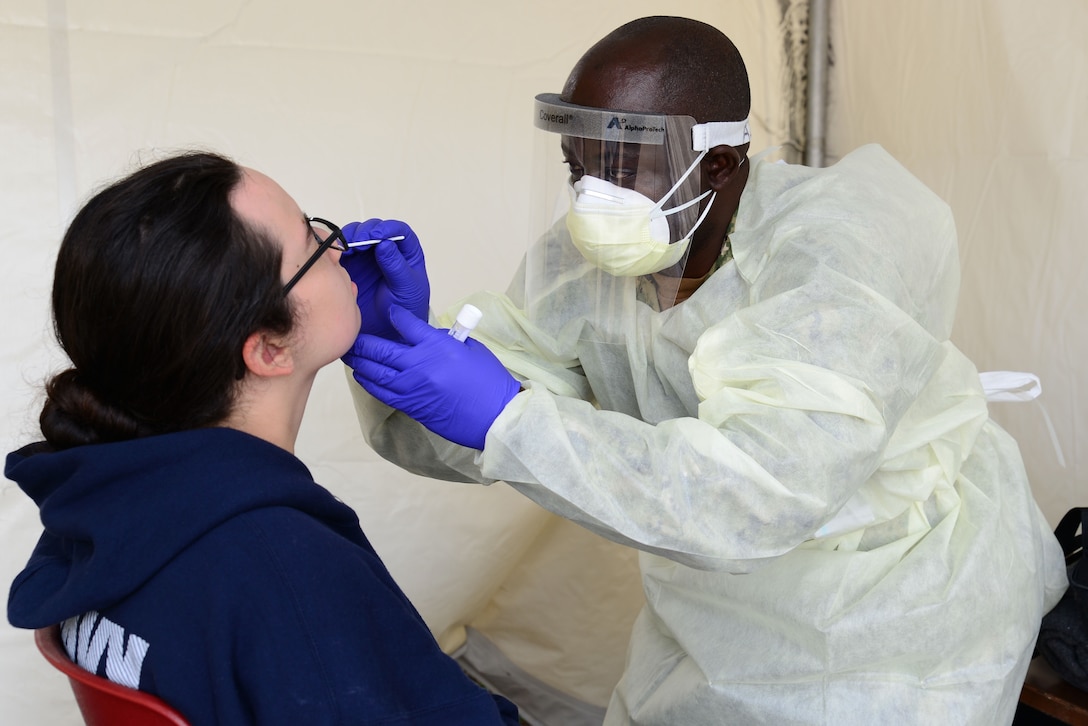 A service member wearing personal protective equipment inserts a swab into a young woman’s nose.