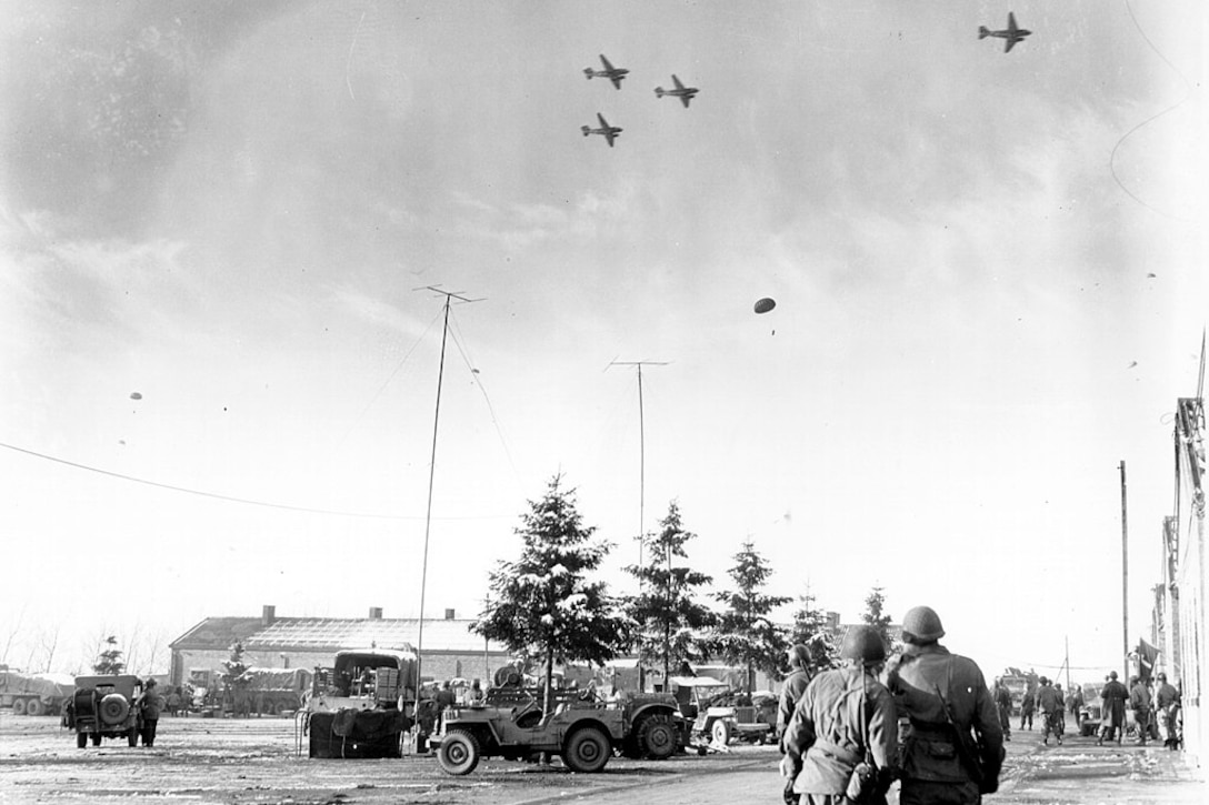 In a historic photo, soldiers on the ground watch as paratroopers descend from the planes flying above them.