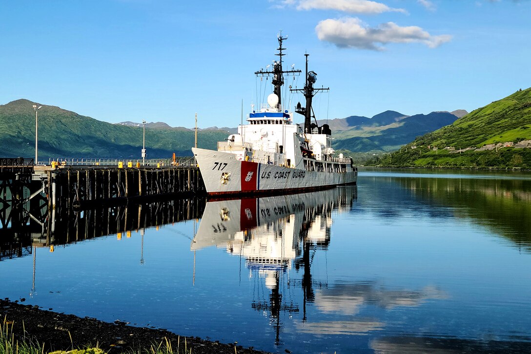 A Coast Guard cutter's reflection is clear in the water as it sits moored at a pier.