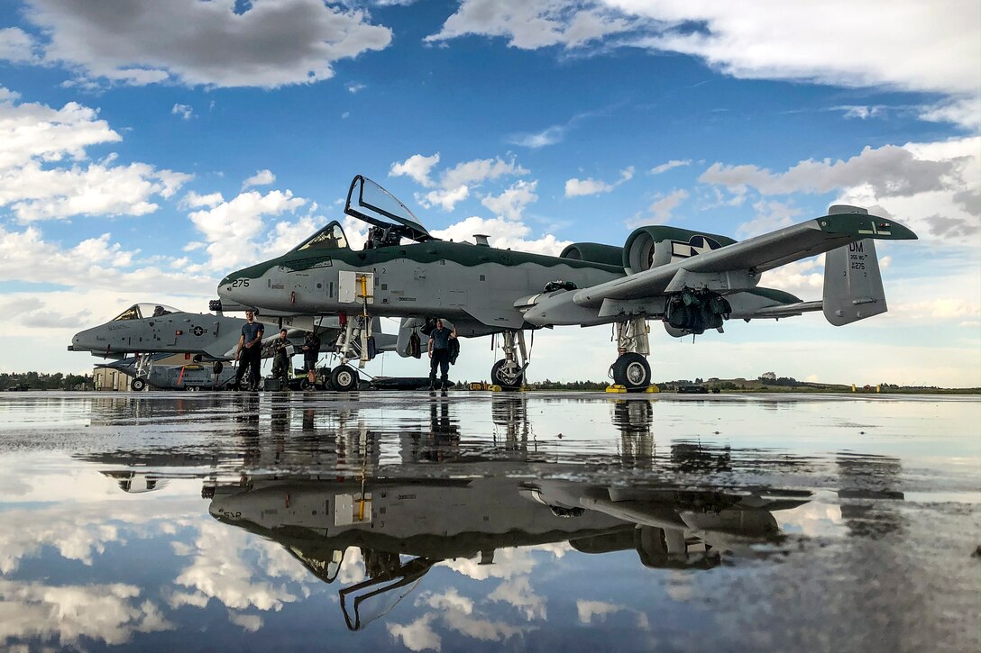 Two aircraft sit on a wet flightline, their reflections visible in a puddle.