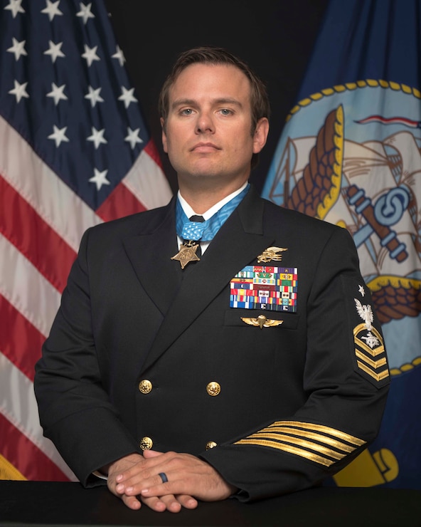 Posing for the picture in his uniform, wearing medal of honor with american flag and navy flag in the background