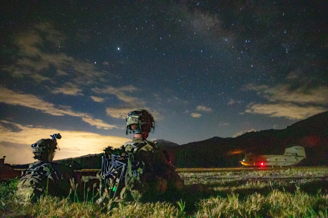Soldiers sit in a large field at night watching a large military helicopter.