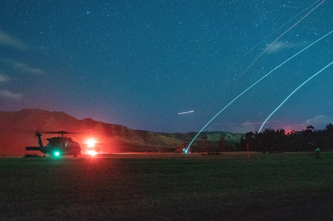 A helicopter sits in a large field with red lights around it while tracers cross a dark sky.