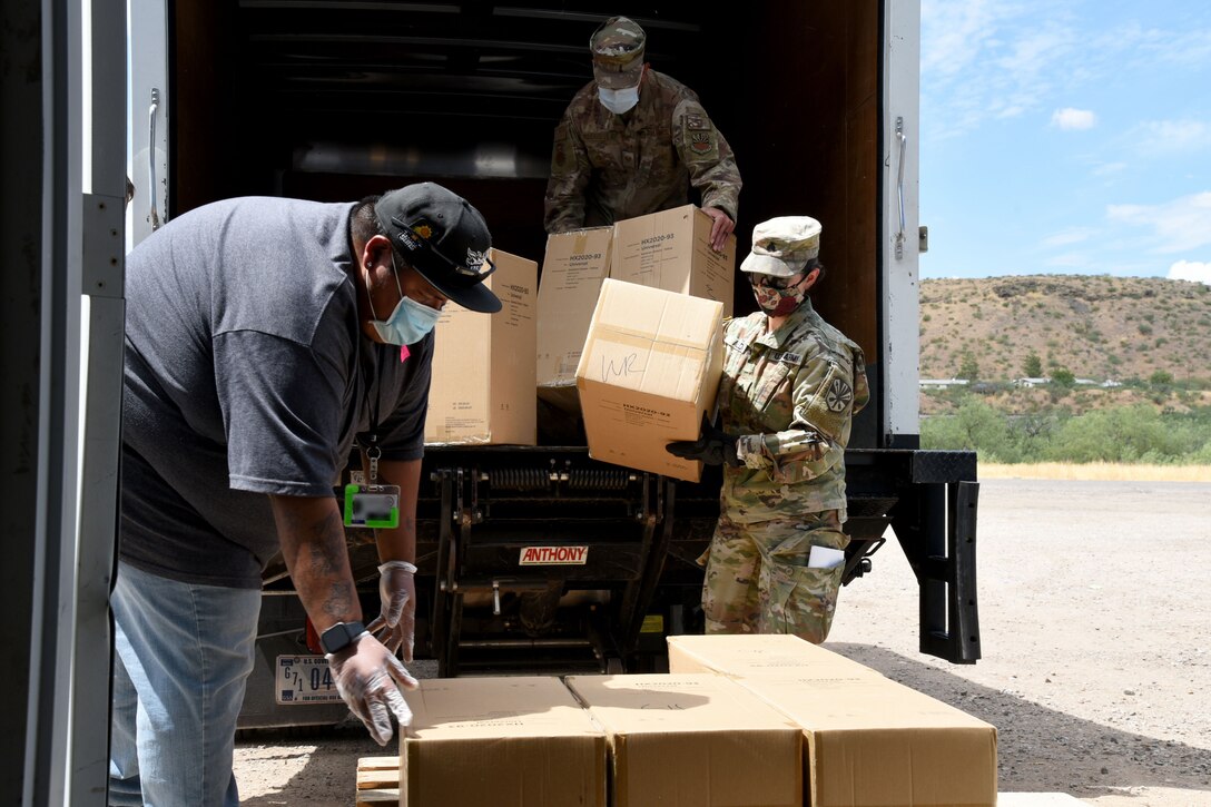 Two guardsmen wearing masks and gloves off-load  boxes of personal protective equipment from a truck. A civilian, also wearing a mask, is in the foreground.