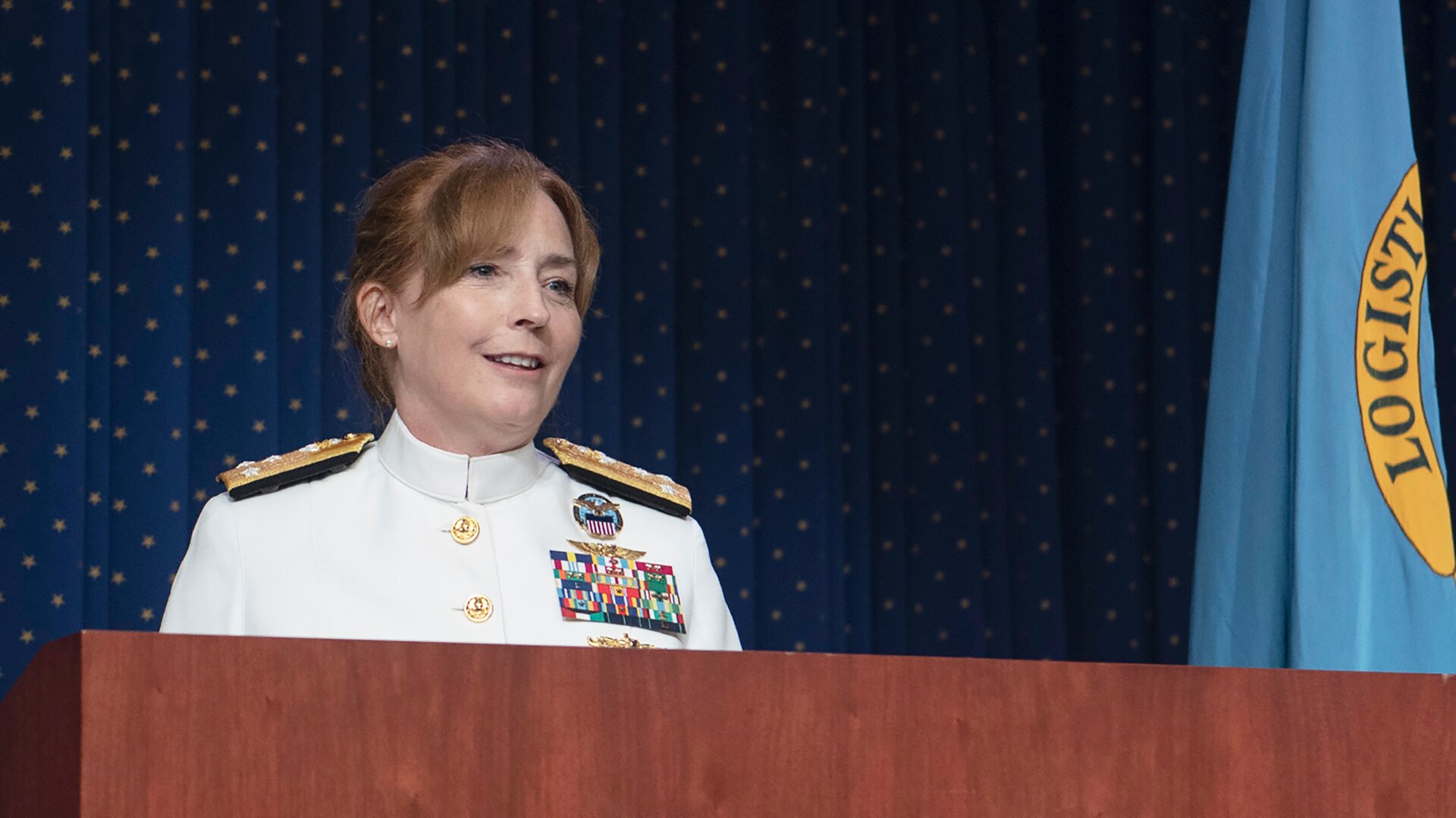 Woman in white Navy dress uniform speaks at a podium with the Defense Logistics Agency flag in the background.