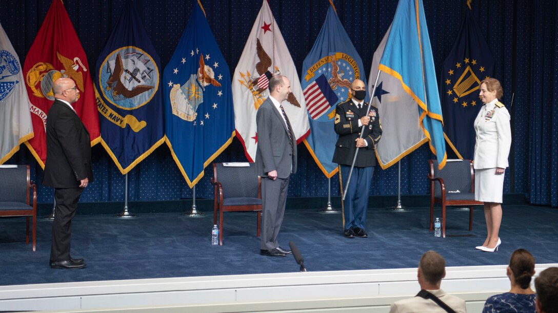 Two men in suits, a uniformed soldier and a woman in a Navy dress uniform stand on a stage with flags representing DLA and the military services.