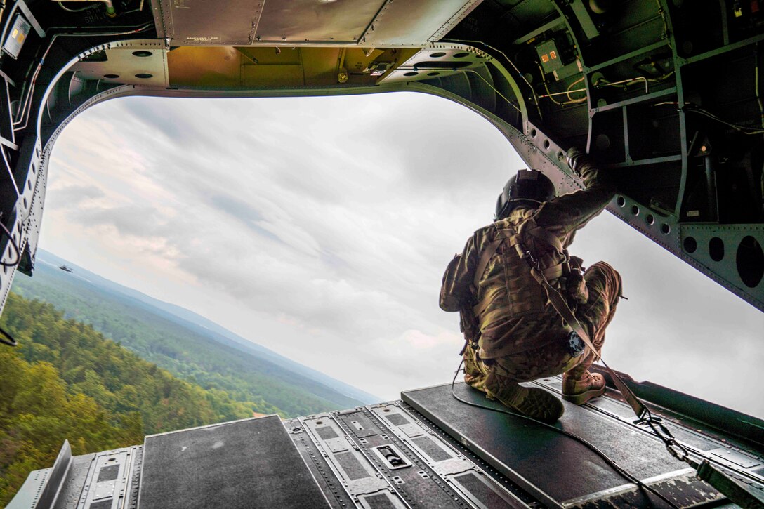 A soldier looks out the back of a military aircraft; a forest seen below.