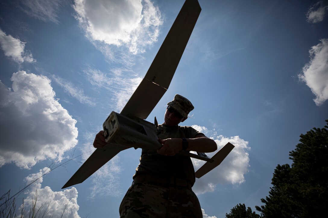 A soldier holds a Raven unmanned vehicle, with a blue cloud-studded sky in the background.
