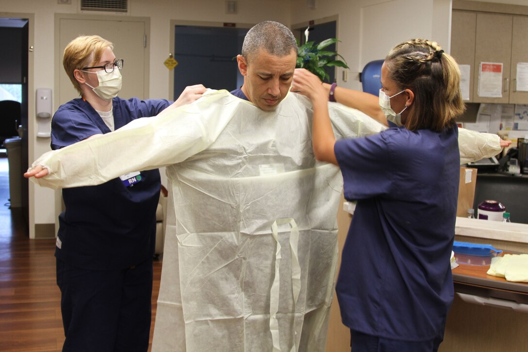 Two Air Force nurses wearing masks help another Airman with putting on personal protective equipment.