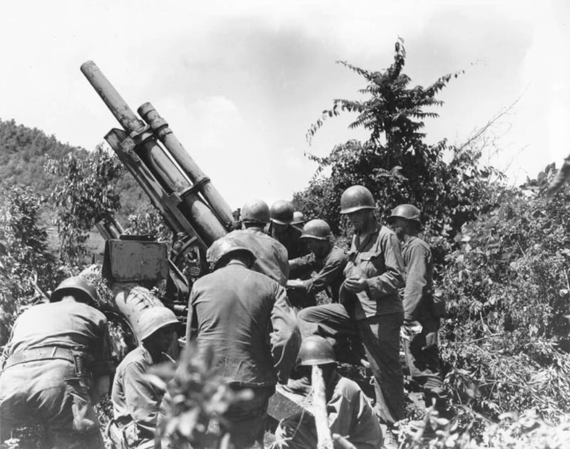 Service members in combat gear surround a gun shrouded in tall shrubs.