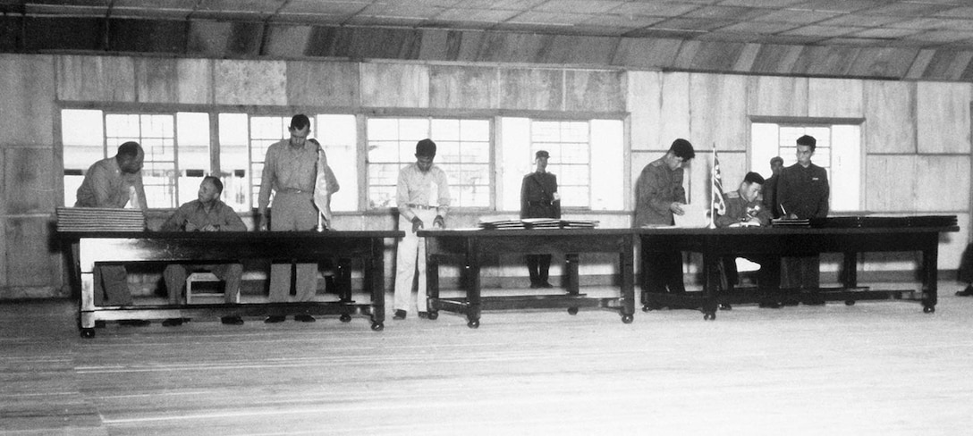 People sit and stand behind three, long tables as guards stand in the background.