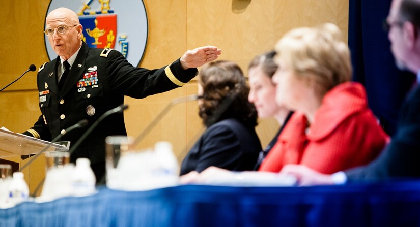 A man wearing a military uniform speaks into a microphone, four people sit at a table in the foreground.