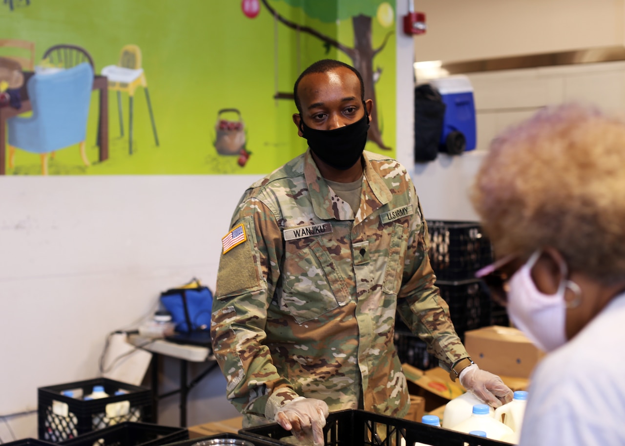 A soldier wearing a face mask and latex gloves helps a visitor at a food bank.