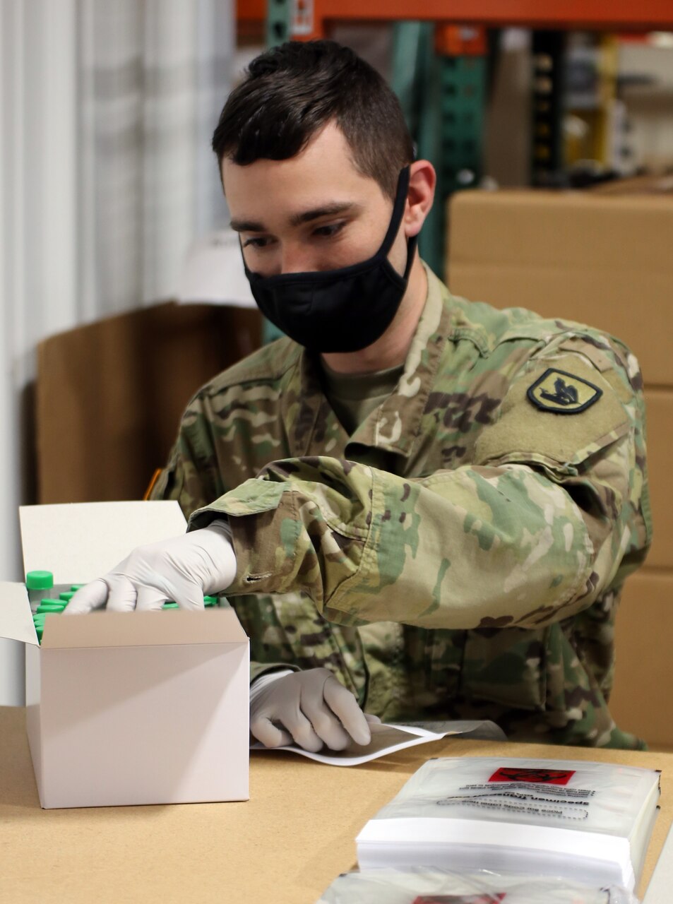 A soldier wearing a face mask reaches into a box of vials while assembling COVID-19 test kits.