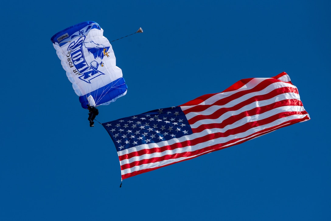 A member of the U.S. Air Force Academy "Wings of Blue" parachute team descends from the sky holding an American flag.