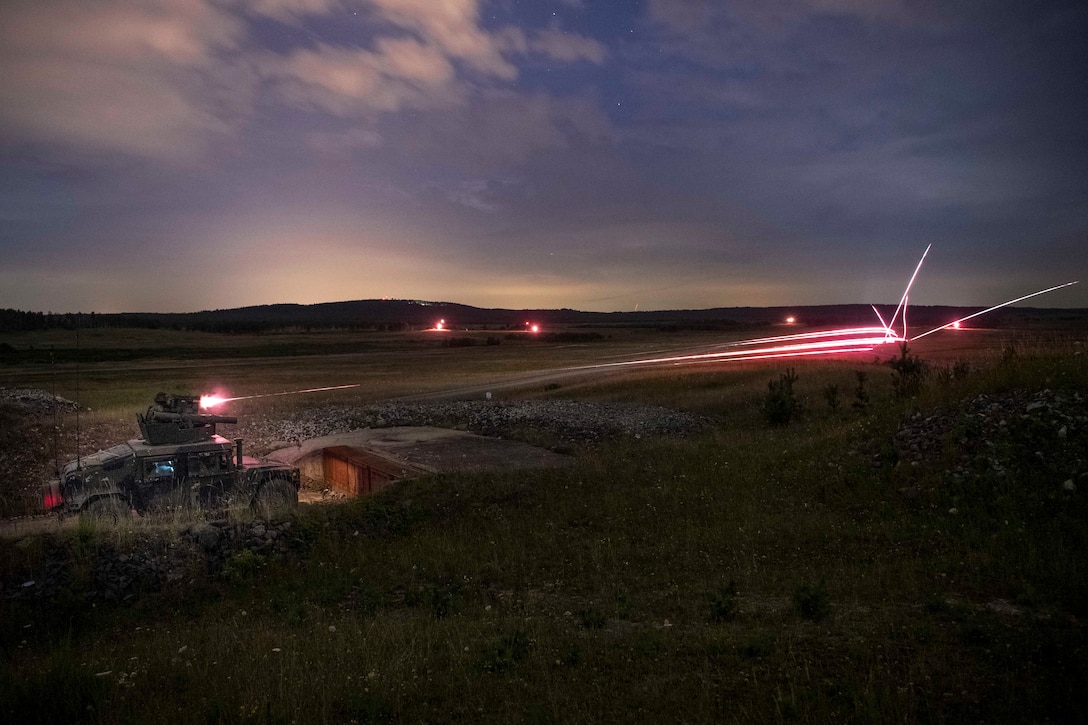 Bright reddish pink lines shoot across a field as a soldier fires a machine gun from a military vehicle at night.