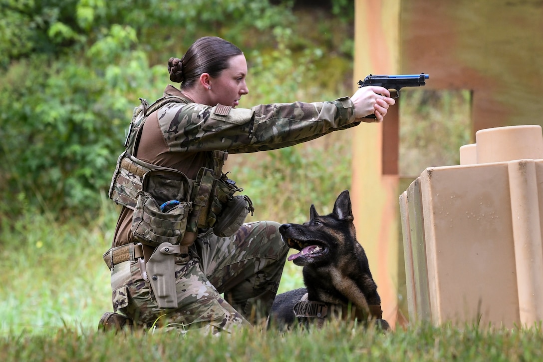 An airman kneeling in grass aims a pistol, as a dog lies next to her and stares attentively.