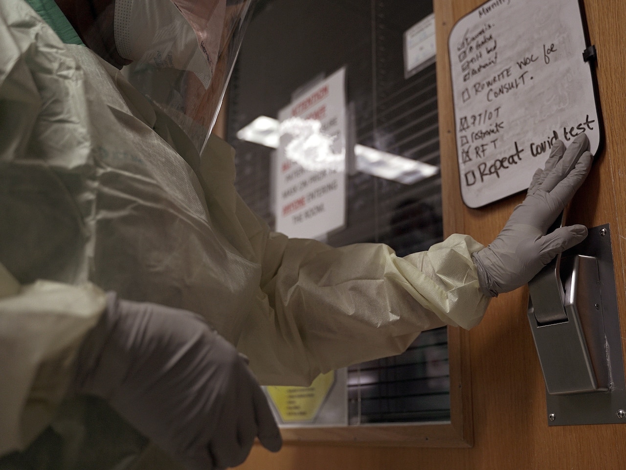 A medical professional wearing protective gear opens a hospital room door with patient notes on the door.