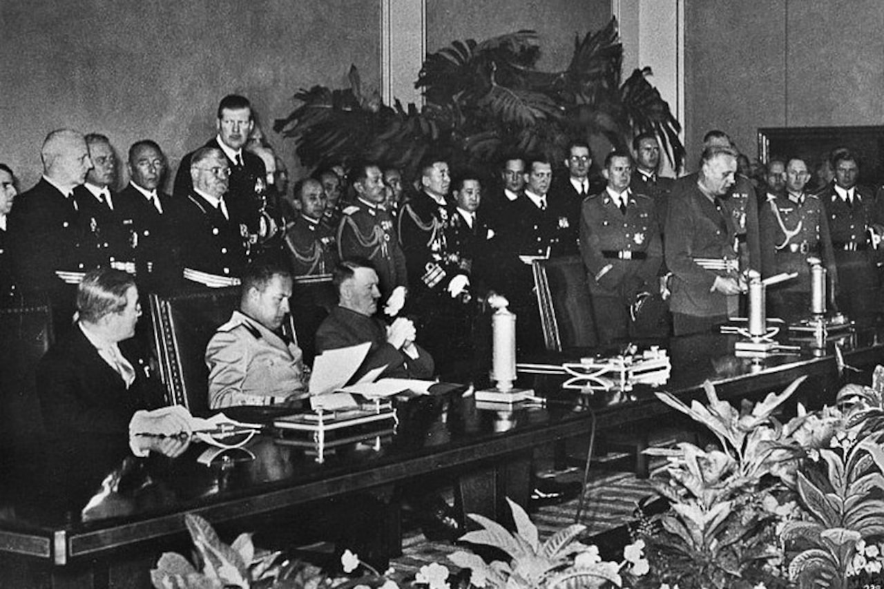 Three men in military uniforms sit behind a long table with lots of people standing behind them.