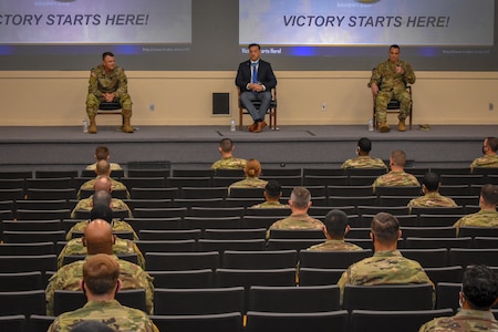 army soldiers sit in a mostly empty theater listening to 3 men speaking on stage.