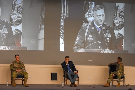 army soldiers sit in a mostly empty theater listening to 3 men speaking on stage.