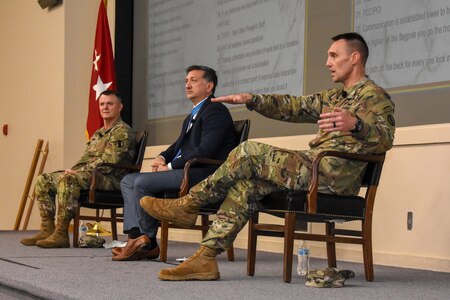 army soldiers sit in a mostly empty theater listening to 3 men speaking on stage.