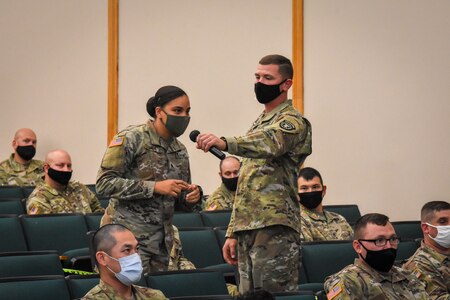 army soldiers sit in a mostly empty theater listening to 3 men speaking on stage.