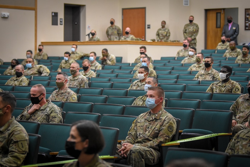 groups of soldiers sit in an mostly empty theater listening to 3 men speak on the stage.