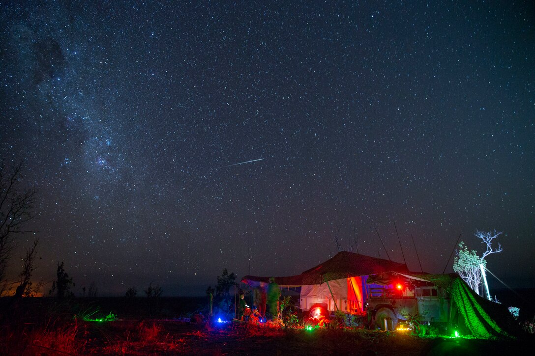 Marines stand near a tent and vehicle under the stars in a field-like area illuminated by green, red and blue lights.