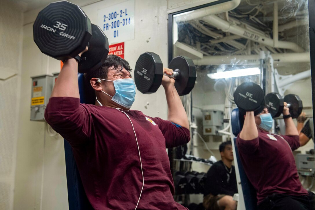 A sailor wearing a mask performs shoulder presses.