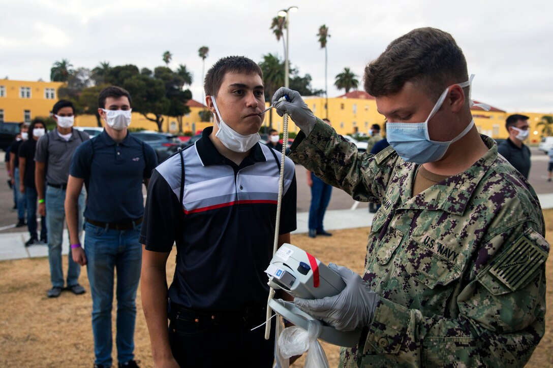 A Marine wearing personal protective equipment takes the temperature of a new recruit who has adjusted his face mask for medical screening while other incoming recruits wearing face masks wait their turn in line.