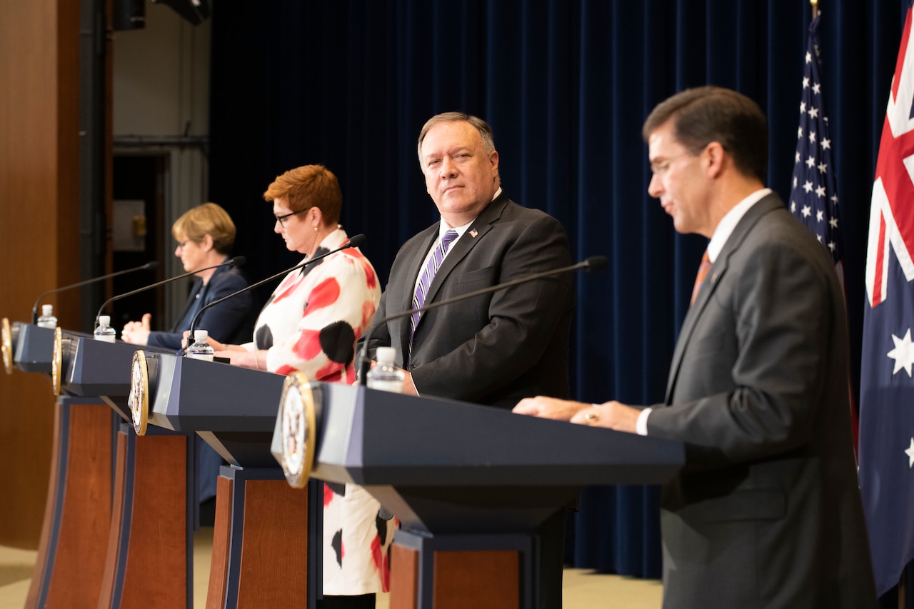 Four people stand on stage at four different podiums.
