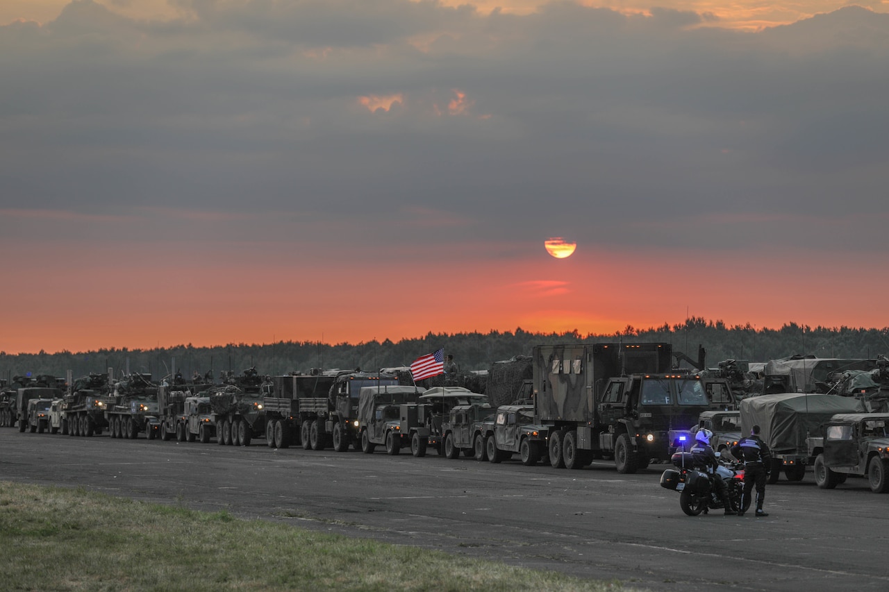 More than a dozen military vehicles line up on a road at dusk.