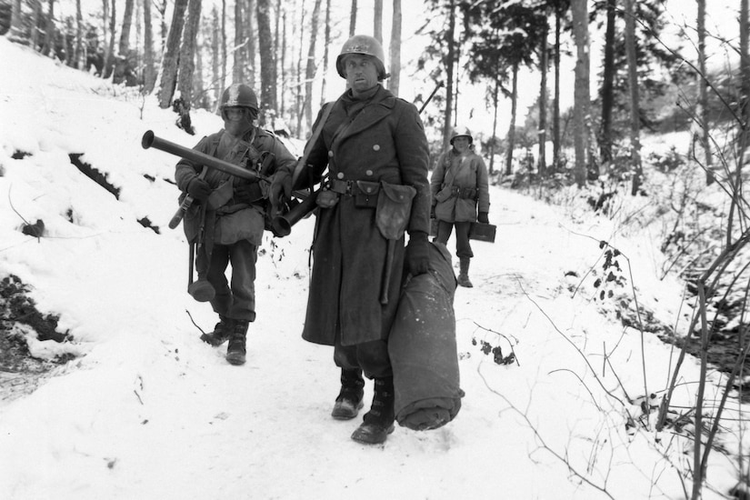 Three soldiers trudge through heavy snow in a forest carrying lots of equipment.