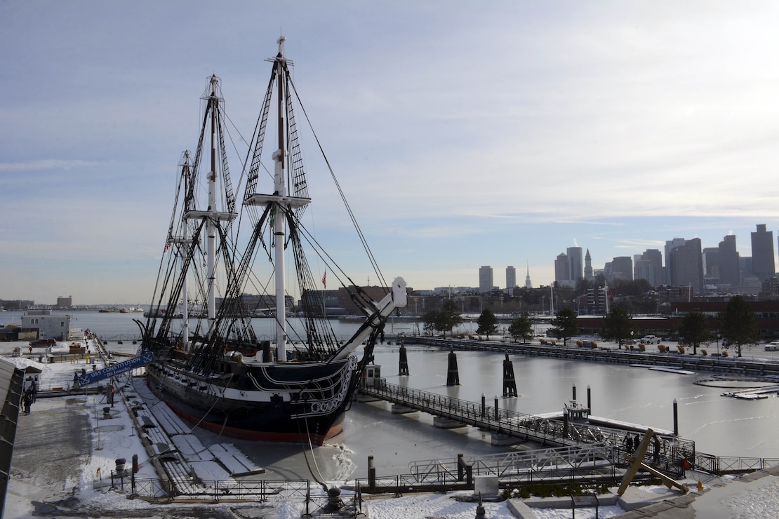 USS Constitution sits moored pier side at Boston Navy Shipyard with boston skyline in the background