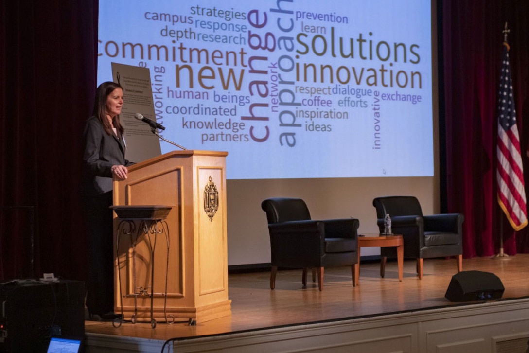 A woman standing at a lectern speaks to a group. Behind her is a word graphic.