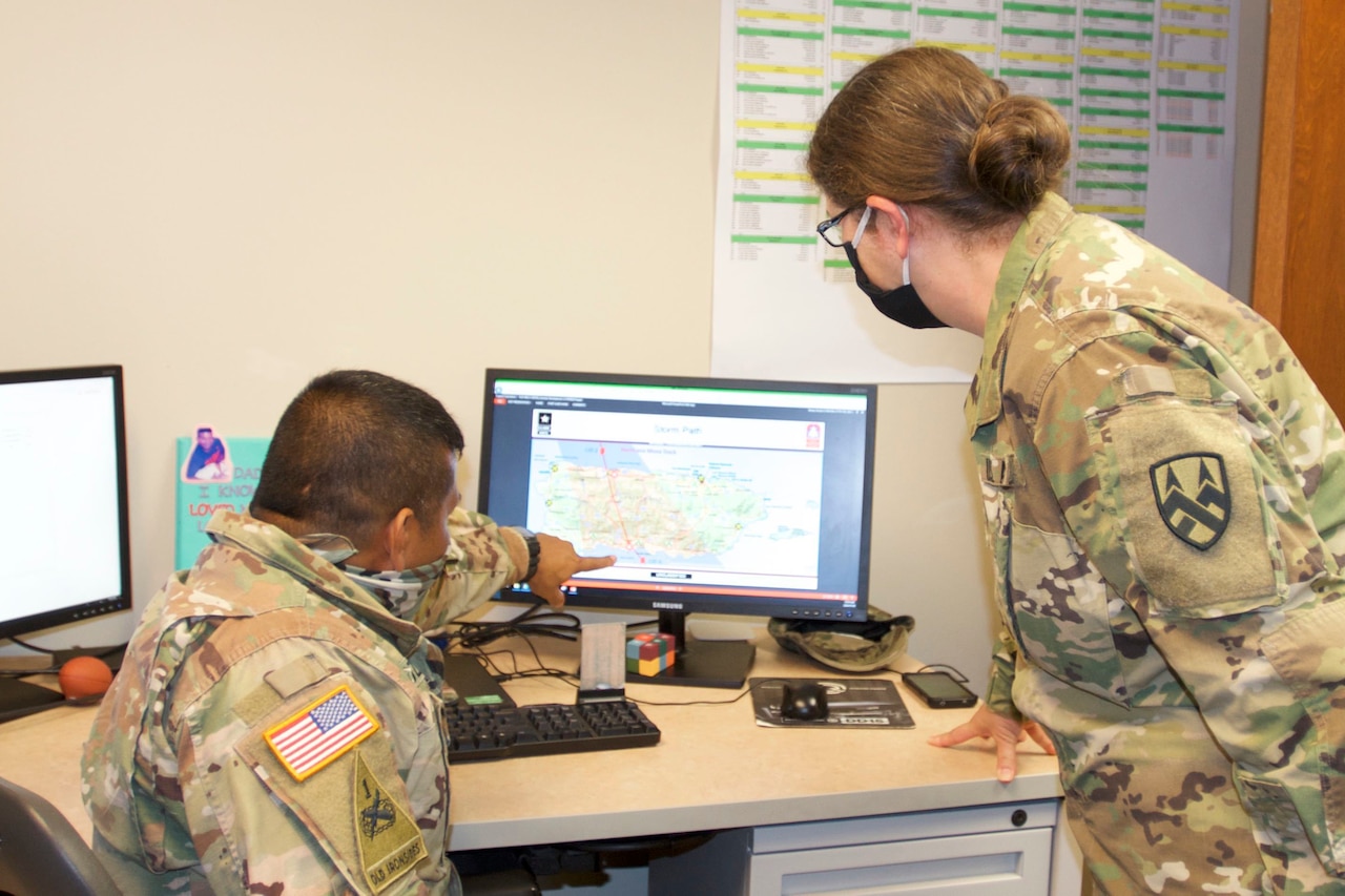 A seated soldier points out a detail on a computer monitor as another soldier standing next to him looks at the monitor. Both are wearing face masks.