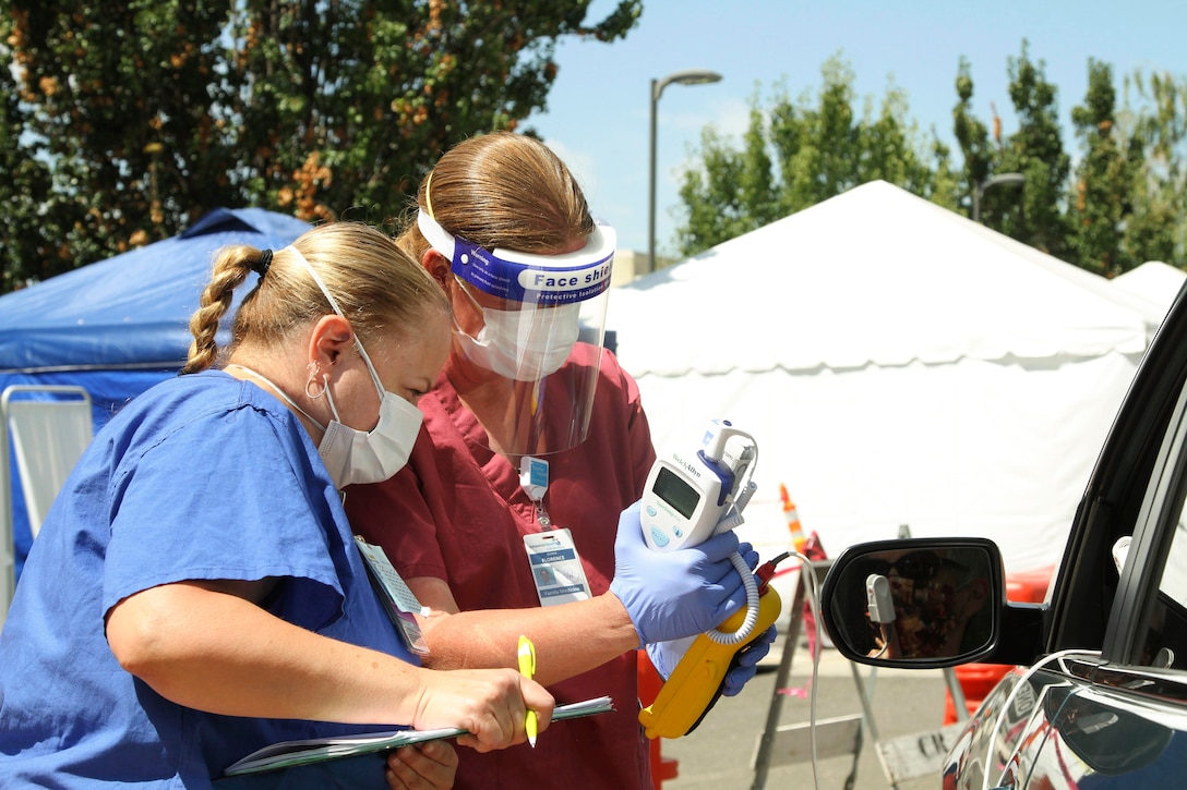 An airman and a nurse wearing protective gear talk to a motorist.