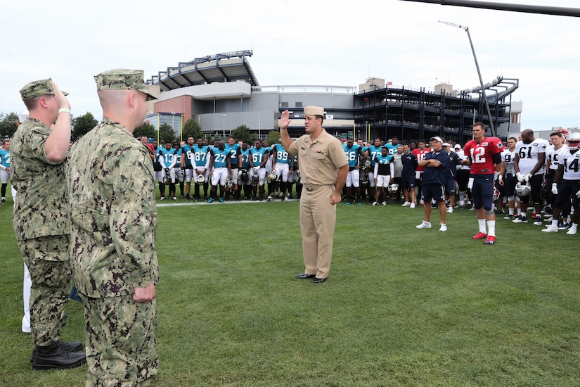 A Navy officer administers an oath to two others in uniform on a football field.