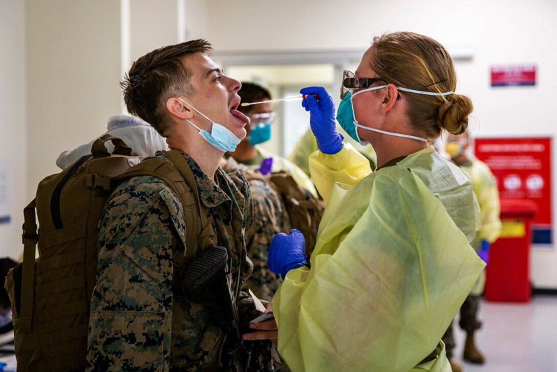 A Marine with a mask below his chin opens his mouth while a person wearing personal protective equipment administers a COVID-19 test.