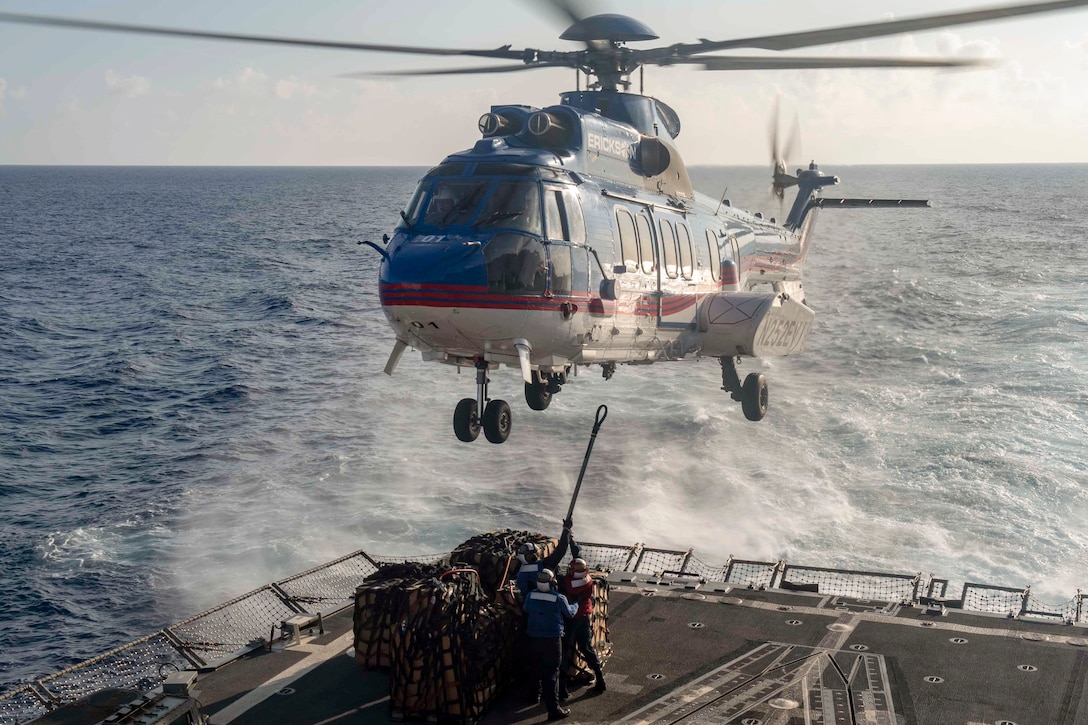 Sailors attach cargo to a helicopter while aboard a ship at sea.